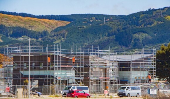 An under-contruction house surrounding by scaffolding, where construction workers stand in high-vis vests